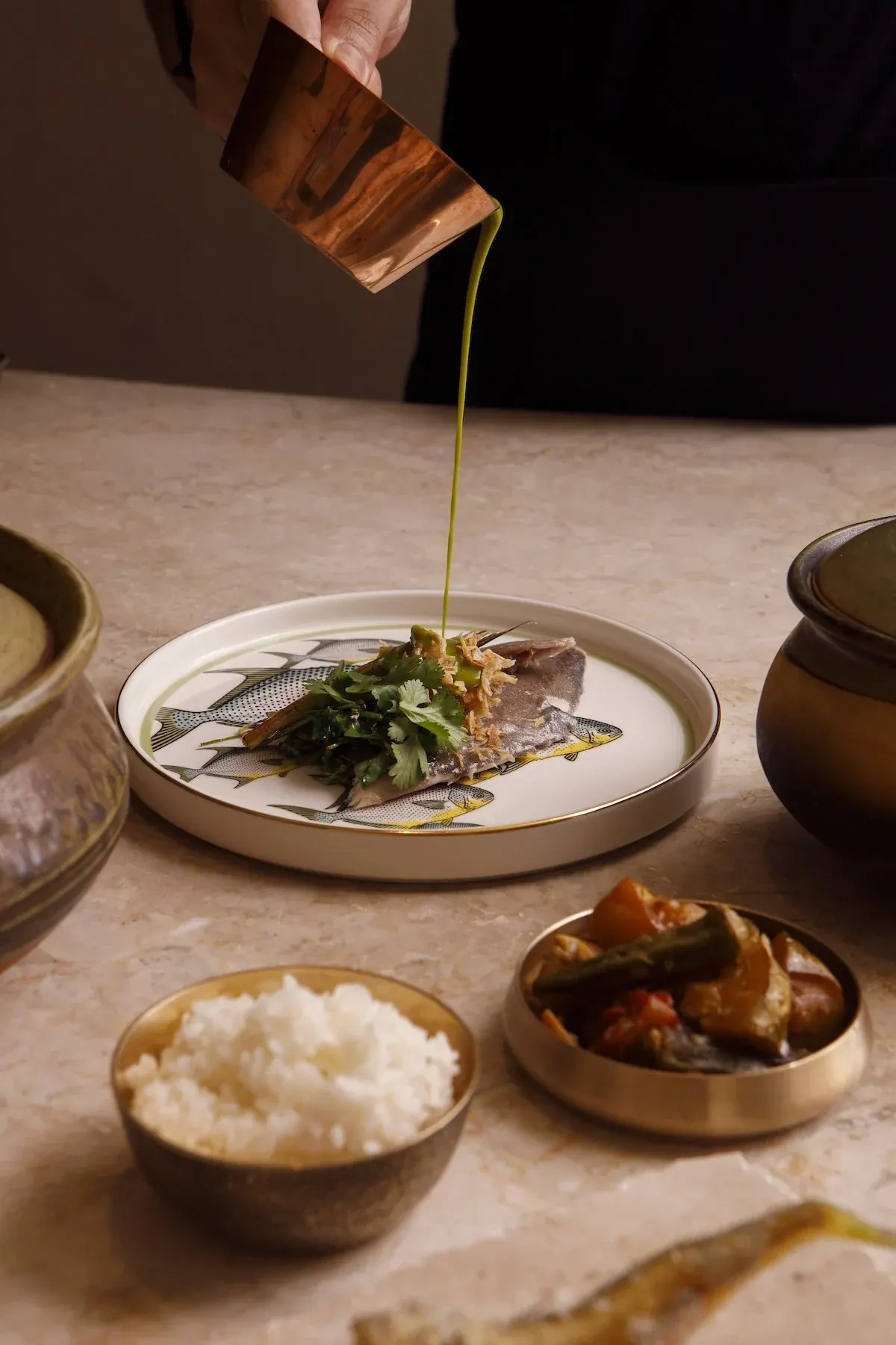 A cook pours sauce over a plated fish beside bowls of rice and side dishes on a stone counter.