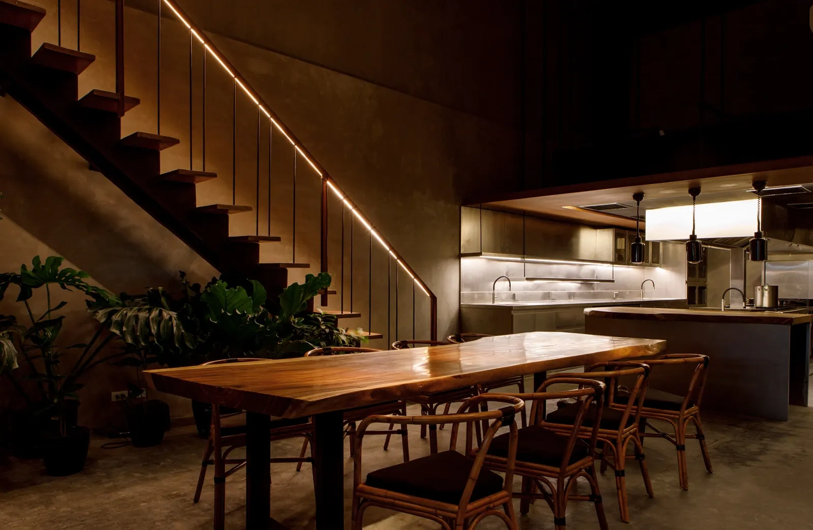A dark wood dining room with a long communal table, a staircase and an open kitchen at Toyo Eatery.