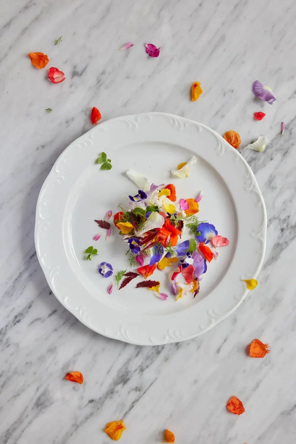 A white plate of flowers and herbs on a marble table scattered with petals at Room4Dessert.
