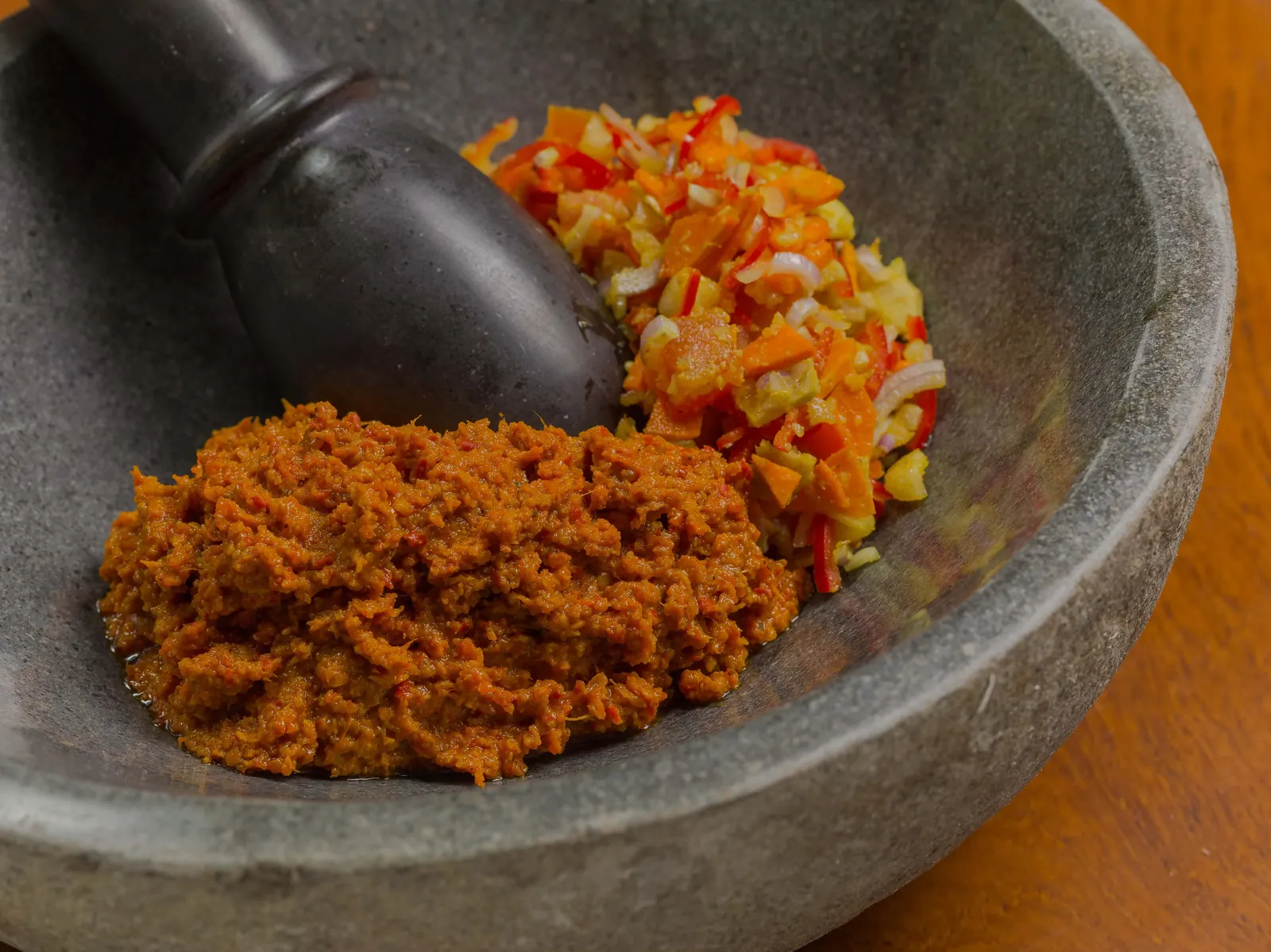 A heap of chopped chilli, shallot and spices beside a ground orange paste in a grey stone mortar with a pestle.