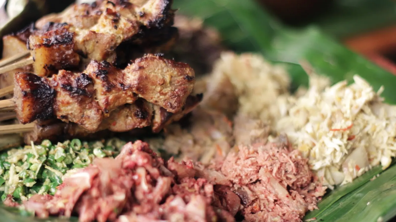 Grilled satay, minced meat dishes, shredded coconut and long beans piled on banana leaves at a megibung in Karangasem.