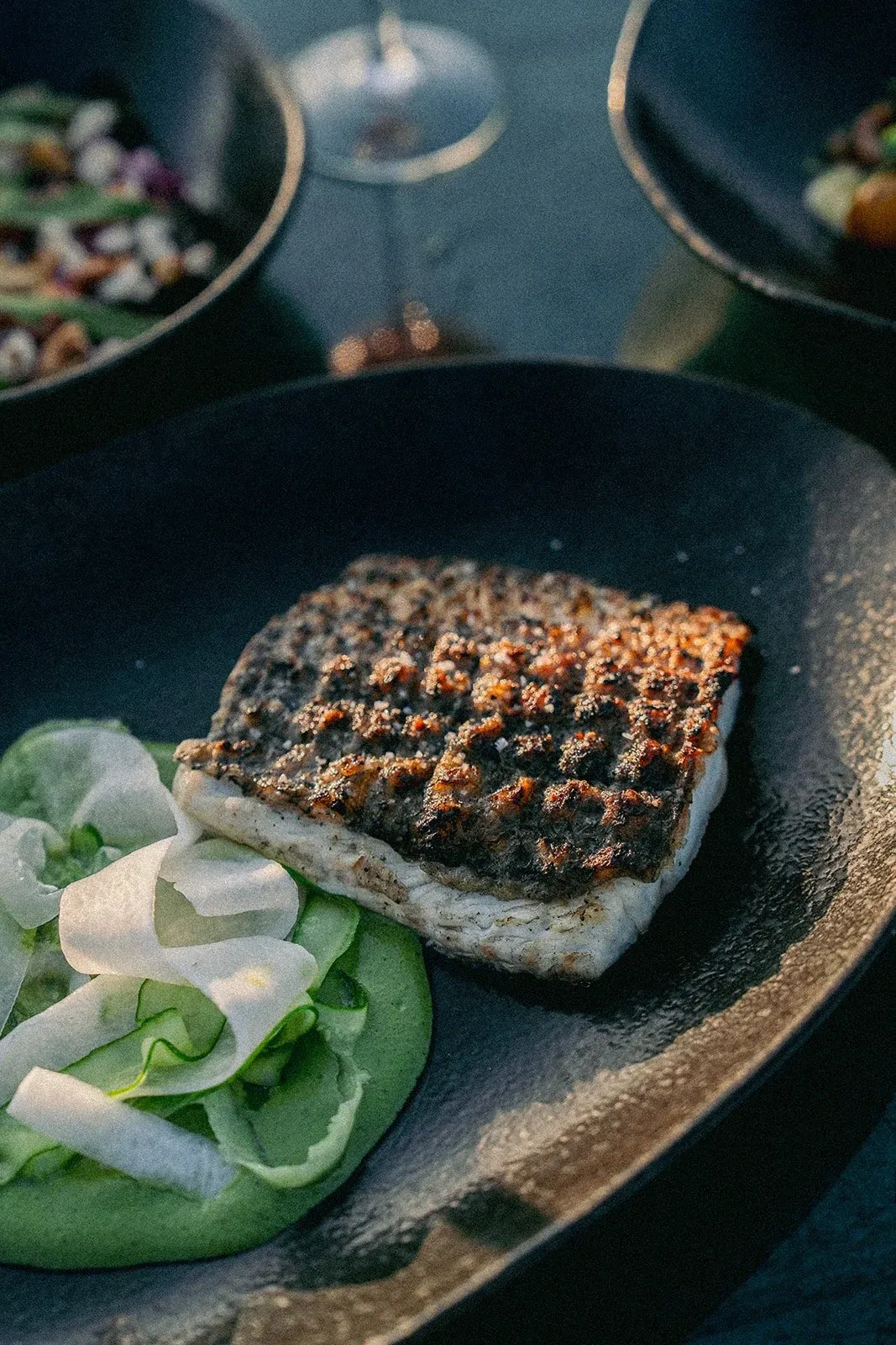 A fillet of grilled fish with a charred crosshatch crust beside shaved cucumber on a green sauce, in a dark bowl at burnt at Ulu Cliffhouse.