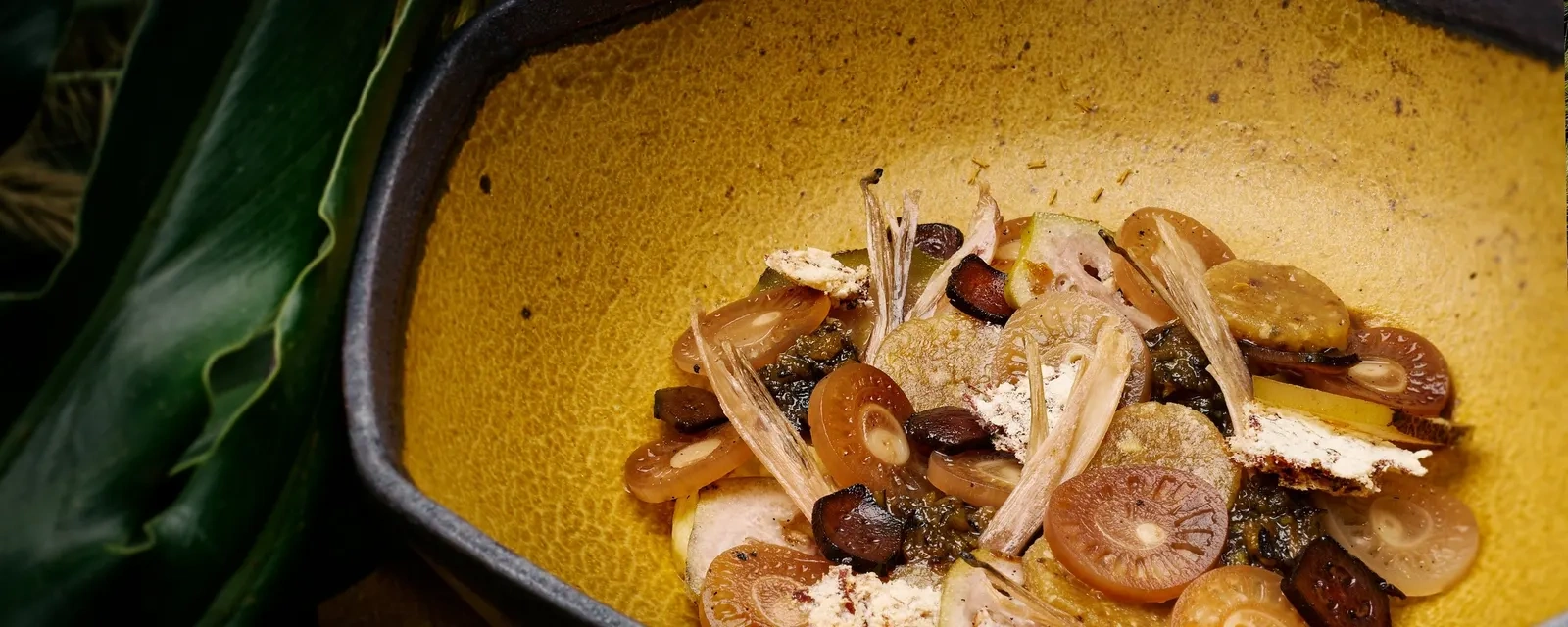 A dish of sliced mushrooms, vegetables and shaved leaves in a mustard-yellow ceramic bowl beside a green leaf at Locavore NXT.